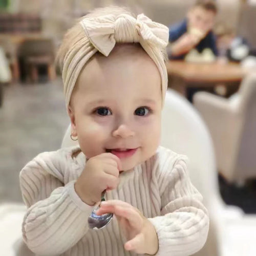 Baby wearing a beige headband with a bow, sitting in a high chair indoors.